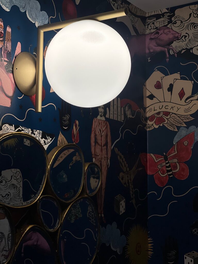 Flos IC Lights W wall sconce, an opal glass globe balanced on a brass arm, against dark illustrated wallpaper