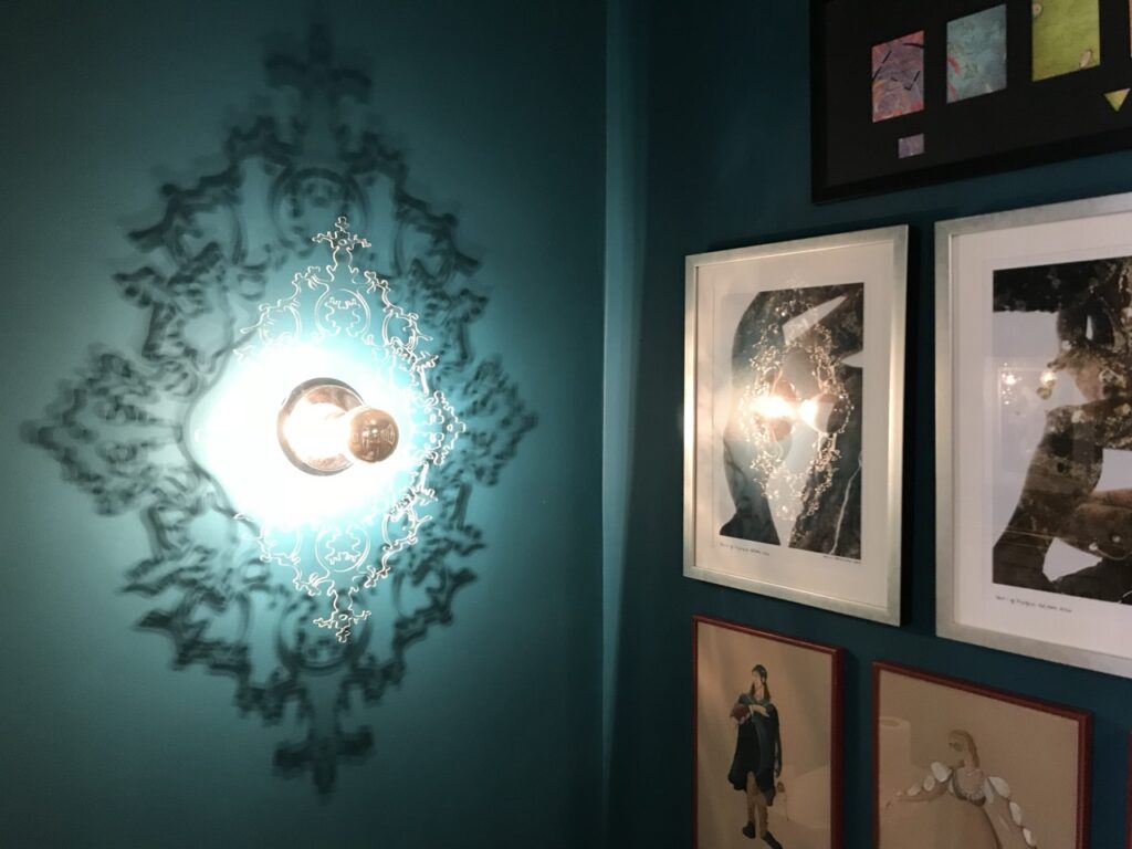 Eurofase Royal wall sconce with a laser-cut baroque frame casting an ornate shadow pattern on a teal wall beside framed artwork