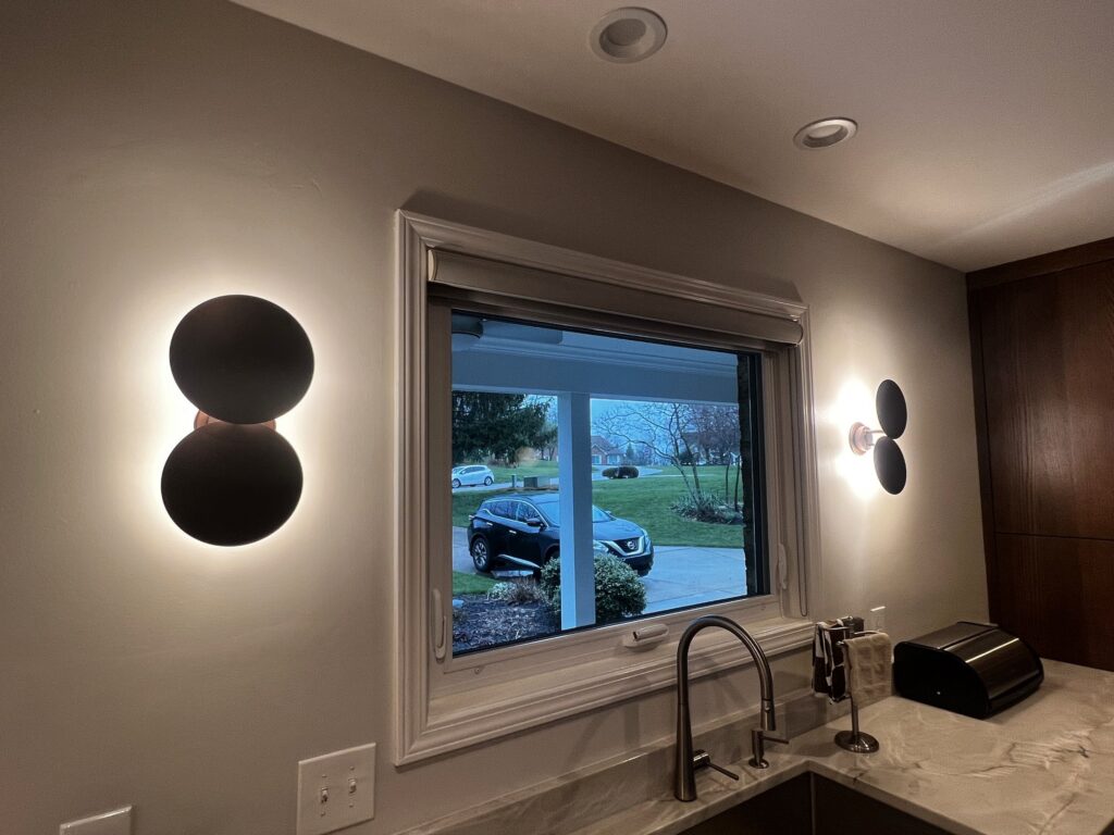 Pair of black double-disc wall sconces with warm halos on either side of a kitchen window above the sink in a Cincinnati-area home