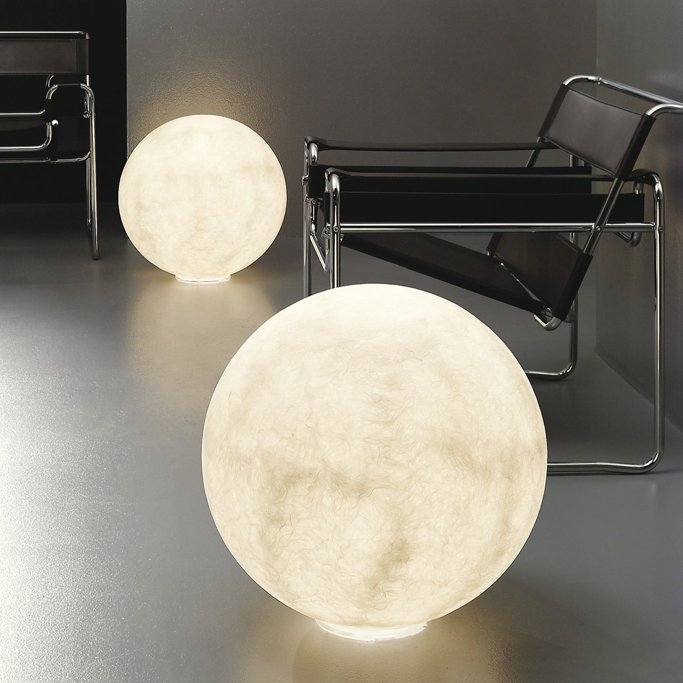 Two Moon lamps in Nebulite, large and small, glowing on the floor beside a lounge chair